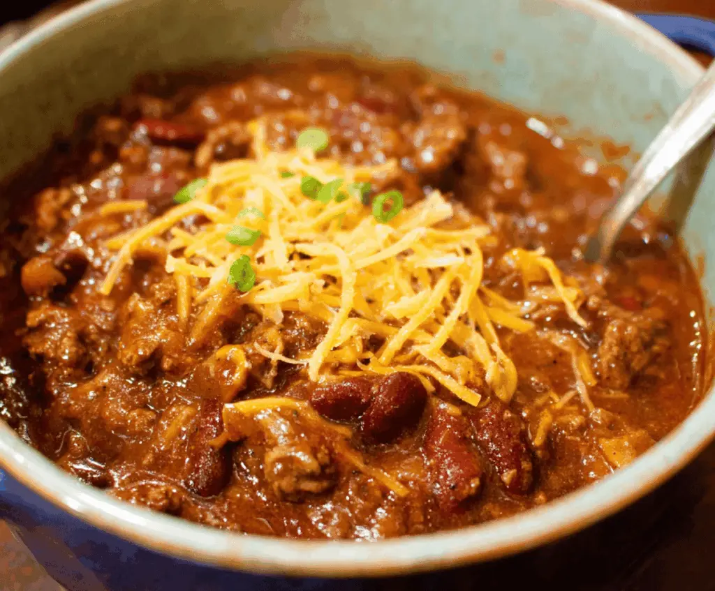 A steaming bowl of The Famous Pioneer Woman Chili topped with shredded cheese, fresh herbs, and sour cream, served in a rustic bowl with a spoon.