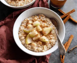 A bowl of warm spiced pear oatmeal topped with cinnamon and chopped nuts, served with fresh pear slices on the side.